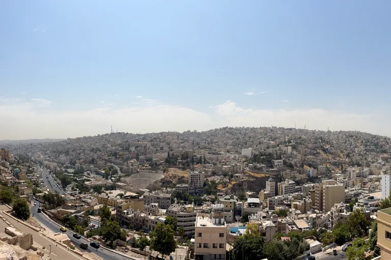 The valley of downtown Amman seen from the Citadel, the curved seats of the Roman Theatre in the middle distance, streets and pale houses filling the valley and covering the hills opposite under a wide blue sky.