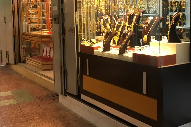 A glass fronted jewellery shop in the Mutrah souq, its lit window hung with gold necklaces on dark display busts, beside an open door and the tiled floor of the covered lane.
