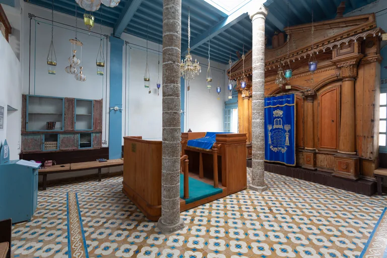 The prayer hall of Slat Lkahal, with a tall carved wooden ark, a blue Torah curtain, a wooden reading desk between two stone columns and a patterned tiled floor.