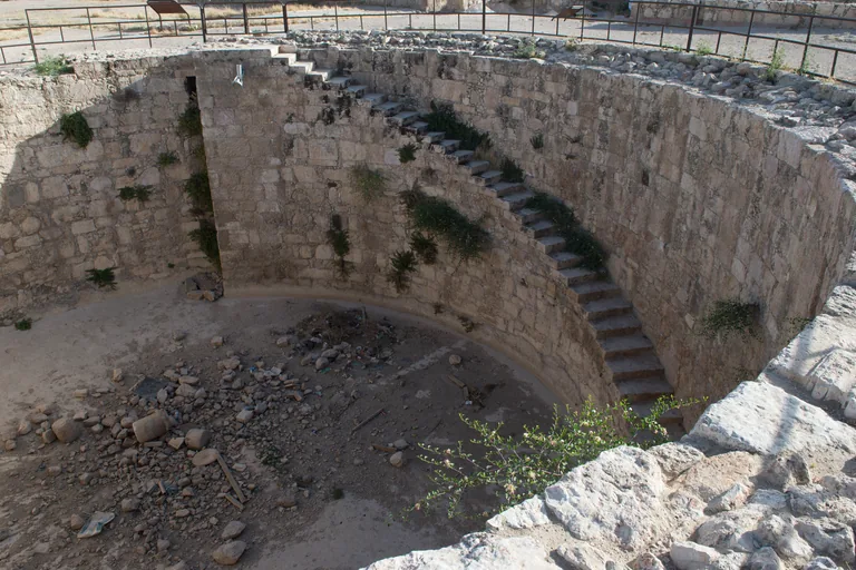 The Umayyad cistern on the Citadel, a deep round stone pit with a stair running down its curved wall, plants growing from the joints and stones scattered on its floor, with a metal railing around the rim.