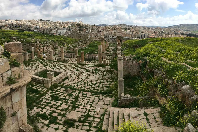 The paved Fountain Court beside the cathedral at Jerash, grass growing between the paving stones and broken columns around it, with a stone stair at the lower right and the modern town on the hills beyond.