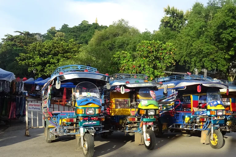 Brightly painted tuk tuks parked in a row on the main street of Luang Prabang, with market stall awnings and racks of clothes at the left, a no parking sign, and a wooded hill topped by a small gilded spire rising above the trees.