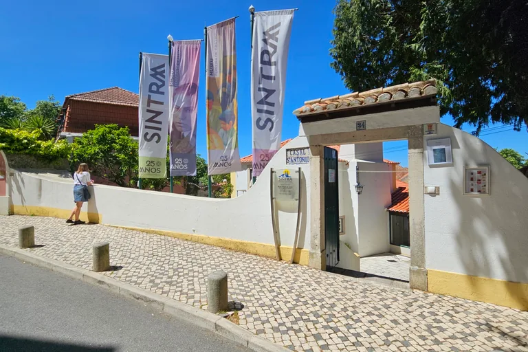 The street gate of the Ferreira de Castro Museum, a white wall with yellow trim and a tiled roof over the entrance, with banners on poles beside it.