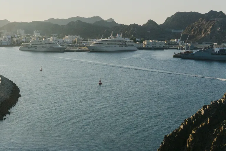The bay of Mutrah seen from a rocky point, calm water with two red buoys leading to large white ships and a grey vessel at the port quays, with the white town and dark mountains behind.