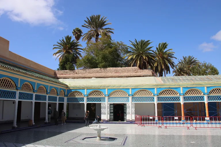 The tiled courtyard of the Bahia Palace with a small marble fountain, blue painted arcades with lattice arches and palm trees behind, part of the arcade under scaffolding and protective sheeting.