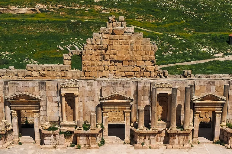 The stage building of the South Theatre at Jerash seen from the seats, its front of columns, pedimented doorways and niches above the stage floor, with a ruined block wall behind and a green hillside beyond.
