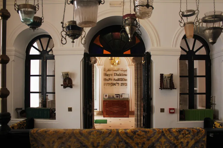 Inside Bayt Dakira, hanging lamps before tall arched windows and an open doorway to a room where the museum's name is written in Arabic, Latin, Tifinagh and Hebrew letters.