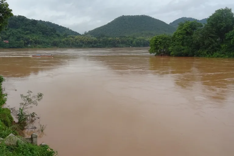 The wide brown water where the Nam Khan meets the Mekong at the tip of the peninsula of Luang Prabang, with a long boat crossing, a wooded point at the right and green hills under a grey sky.