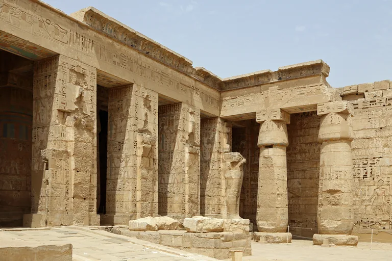 A corner of the second court at Medinet Habu, square pillars with a broken statue of the king as Osiris and round columns, with painted reliefs on the walls behind.