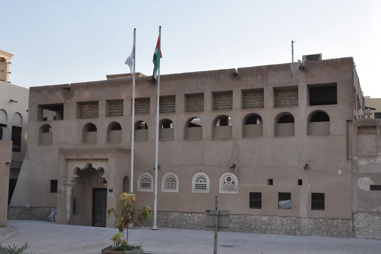 The two storey front of Al Ahmadiya School, plastered walls with rows of arched openings, a cusped arch over the gate and carved round window screens above a base of rough stone, with two flagpoles.