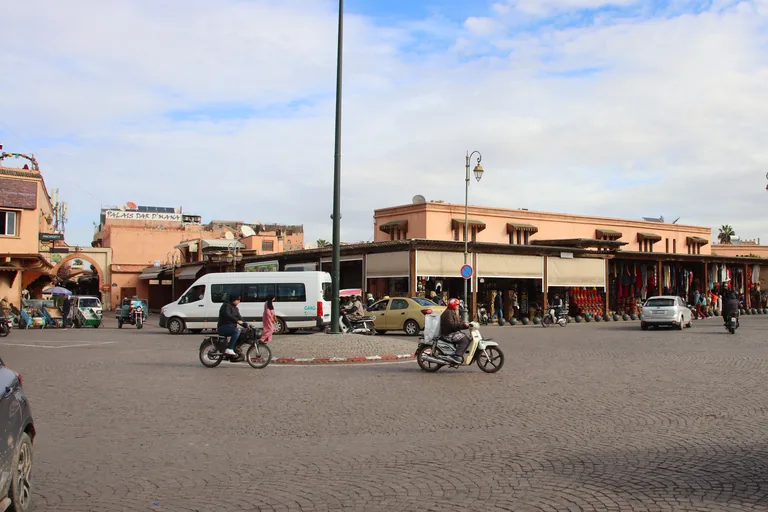 The cobbled Place des Ferblantiers with motorbike riders crossing, a white minibus and a taxi, and shops under awnings along its far side.