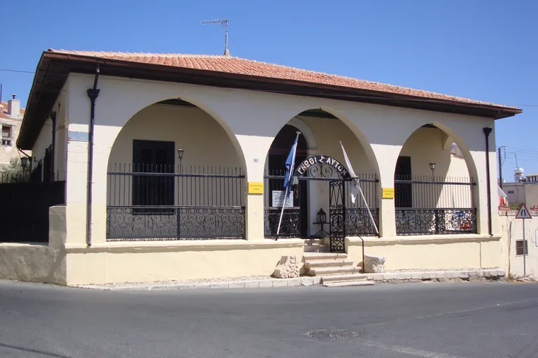 The pale front of the Ethnographic Museum in Paphos, three broad pointed arches behind iron railings with an iron gate under an arch of Greek letters, two flags and a tiled roof above.