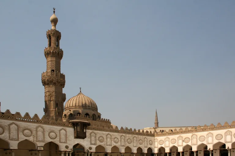 The courtyard of al Azhar, with a carved stone minaret and a ribbed dome rising above white keel arched arcades decorated with carved medallions and crowned with stepped crenellations.