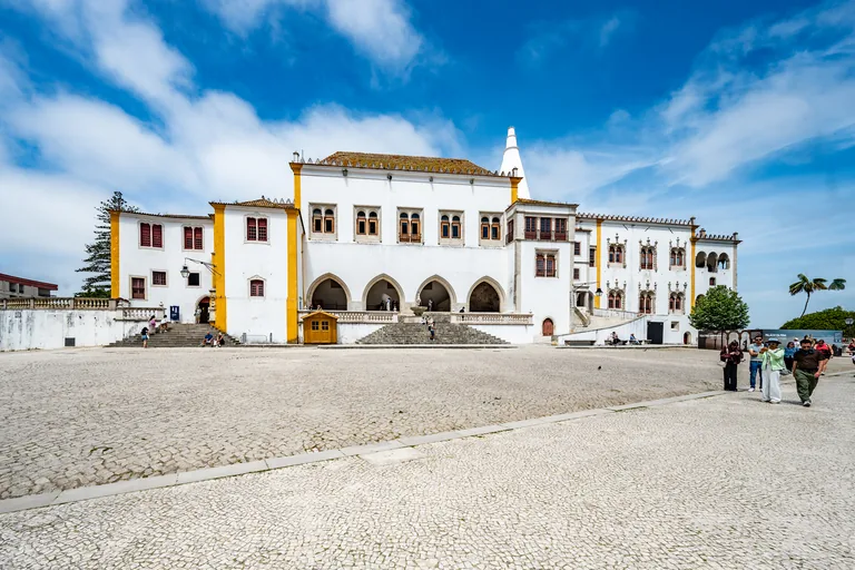 The white front of Sintra National Palace with its arcade, arched windows and a conical kitchen chimney, seen across the wide cobbled square.