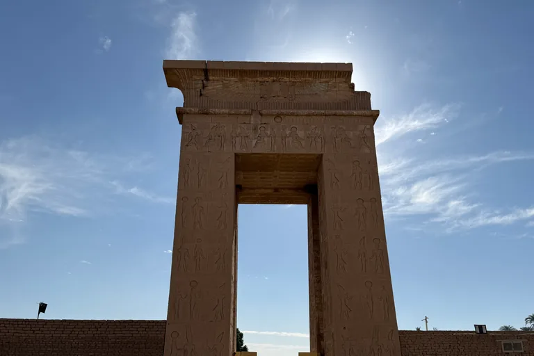 Bab el Amara, the tall Ptolemaic stone gateway carved with reliefs of the king and the gods, standing alone against a blue sky with the sun behind its cornice and a mud brick enclosure wall on either side.