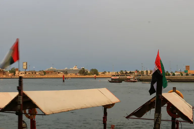 The canopied roofs of moored abras and two flags at the Bur Dubai quay, with the creek, small boats and the low far bank beyond under a grey sky.