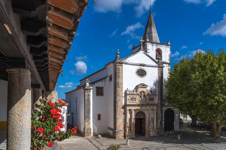 The white front of the Church of Santa Maria, with its carved Renaissance portal and a bell tower with a pointed spire, seen from under a wooden arcade beside red geraniums.
