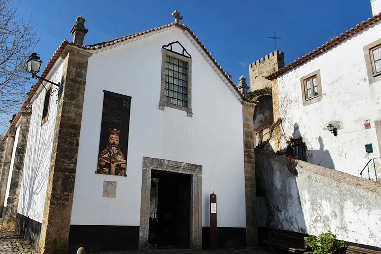 The whitewashed front of the Church of São João Baptista, with a stone framed doorway, a barred window and an exhibition banner, and a castle tower behind.