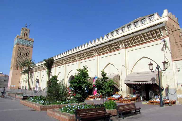The long cream wall of the Kasbah Mosque with pointed arches and its tiled minaret at the far corner, palm trees and flower beds in front, and pottery stalls under awnings along the wall.
