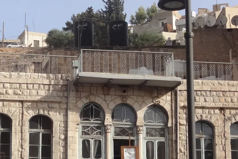 The upper floor of the Duke's Diwan on King Faisal Street in Amman, a stone townhouse with three tall arched windows and a clock in the middle one, under a balcony, with water tanks and trees on the slope above.