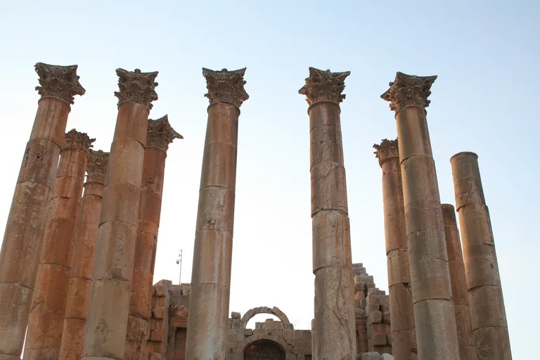 Columns of the temple of Artemis at Jerash with their carved Corinthian capitals against a pale sky, and the arched doorway of the inner hall between them.