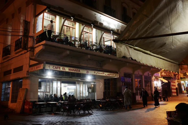 The Petit Socco at night, with the lit Café el Manara on the ground floor of the Hotel Fuentes, empty chairs on the terrace and a few people passing in the square.