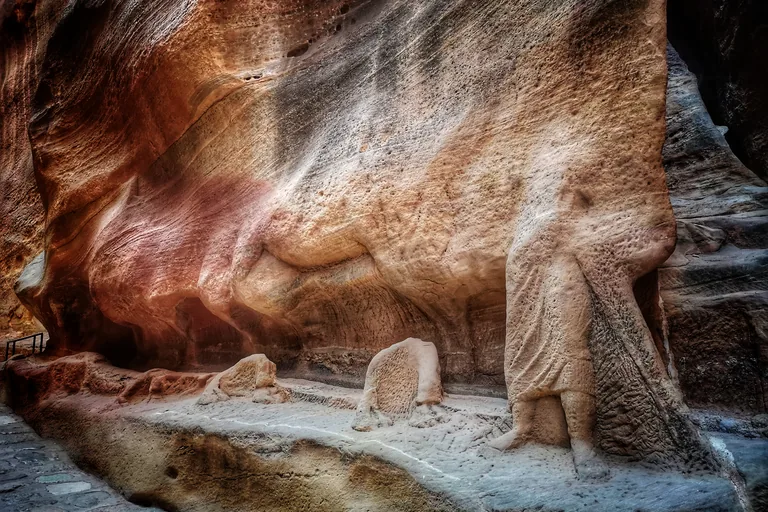 The carved legs and long robe of a drover from the camel caravan relief at the foot of the Siq wall, with worn stone blocks on a ledge beside the paved path.