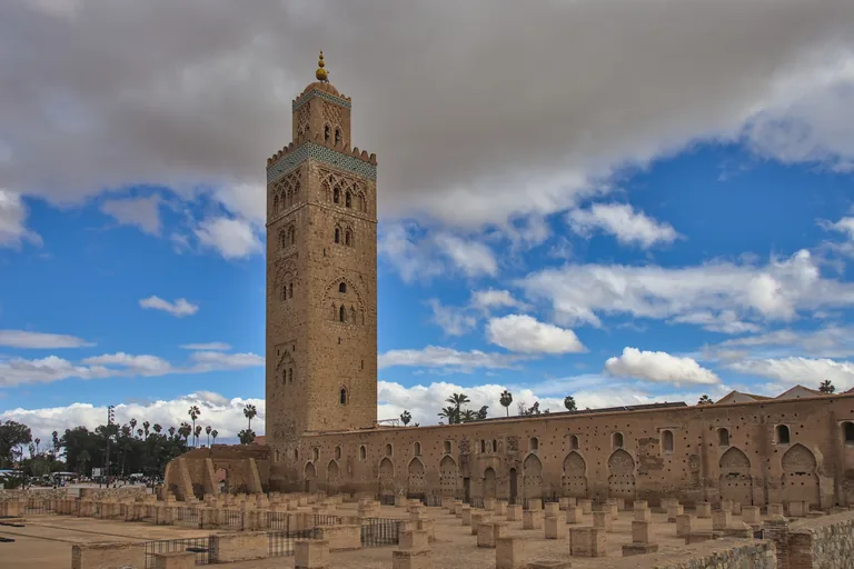 The square stone minaret of the Koutoubia under a cloudy blue sky beside the long arcaded brick wall of the prayer hall, with rows of excavated pier bases of the first mosque in the foreground.