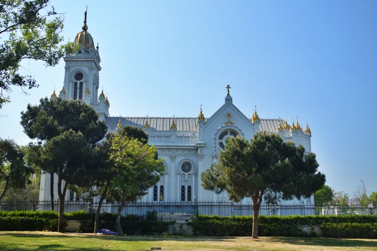 The white cast iron Bulgarian St Stephen Church behind garden railings and pine trees.