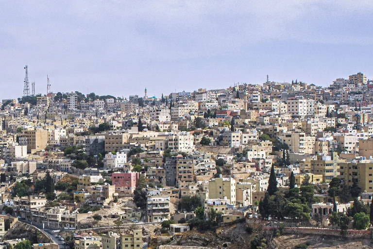 Pale houses and apartment blocks covering the hill across the valley from the Citadel in Amman, with cypress trees among them, a road crossing the lower slope and telecommunication masts on the skyline.