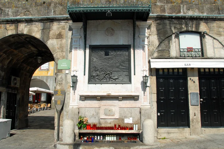 The bronze relief of the Alminhas da Ponte under an iron canopy, with candles on the ledge below.