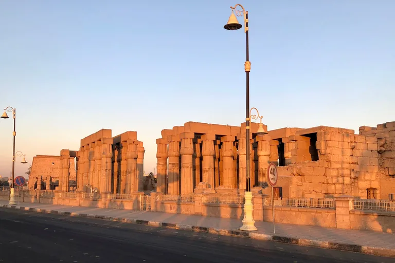 The colonnade and court columns of Luxor Temple glowing in low sun behind a low wall and railings along the Corniche pavement, with the temple pylon in the distance on the left and a street lamp and a speed sign by the road.