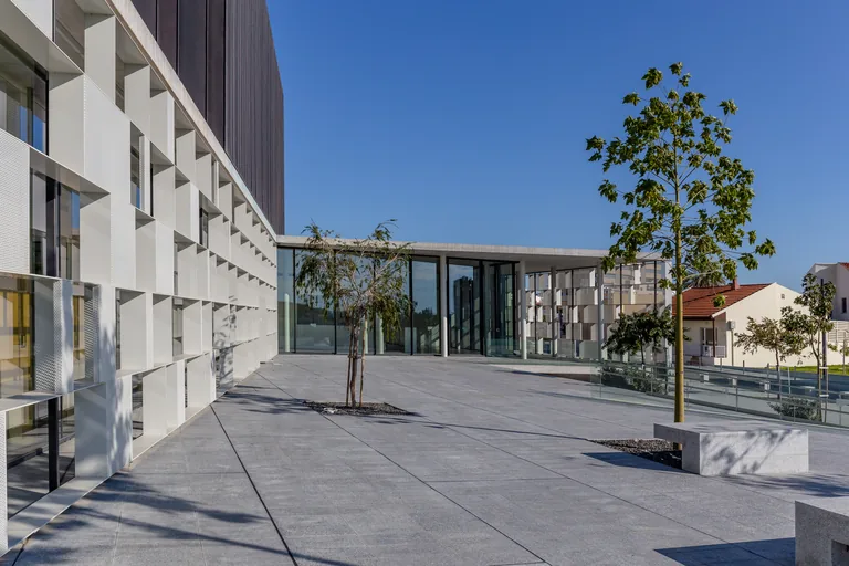The Markideion theatre in Paphos, a long block of white louvred panels with a glass foyer opening onto a wide paved terrace with young trees and a stone bench.