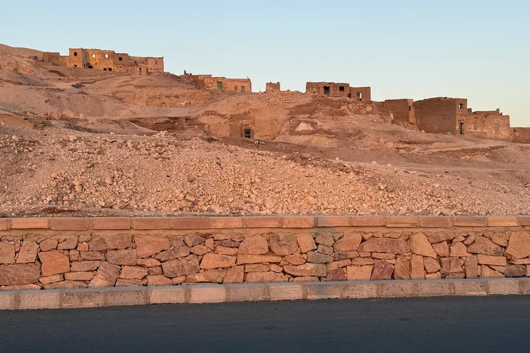 The bare stony slope of Qurnet Murai rising above a stone retaining wall beside the road, with the mudbrick houses of the old village along its crest in evening light.