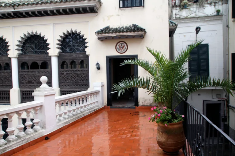 The courtyard terrace of the American Legation, with carved wooden window screens under scalloped arches, the seal of the United States over a doorway and a palm in a large pot.