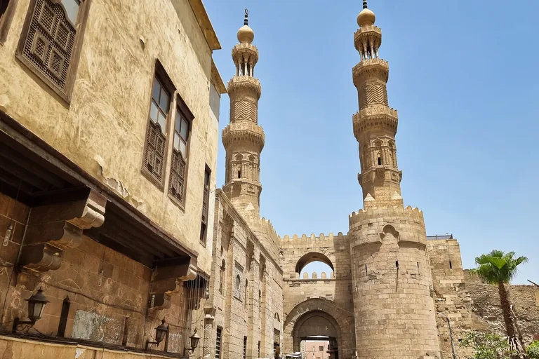 Bab Zuwayla from al Muizz Street on the north, a round crenellated stone tower and arched gateway with two tall carved minarets standing on the towers, an old house with wooden window screens on the left.