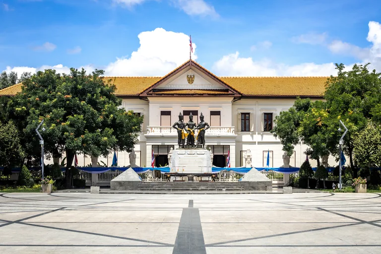 A wide paved square with dark lines crossing it, and three bronze figures standing together on a stone base at its far end, in front of a long white two storey hall with a tiled roof, among trees in the middle of Chiang Mai.
