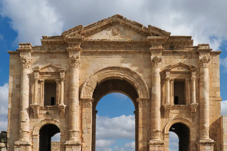 The upper part of Hadrian's Arch at Jerash, a pale stone gateway with a triangular pediment, Corinthian columns on its face, a tall central arch and two smaller arches under framed niches, against a cloudy blue sky.