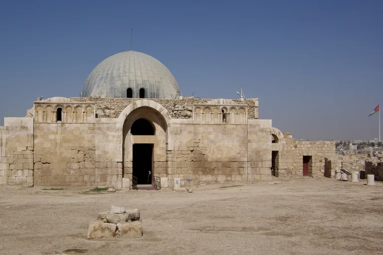 The Umayyad entrance hall on the Citadel, a square stone building with a tall arched doorway, rows of small arches above and a grey ribbed dome, standing on open dusty ground with a flag on a tall pole at the right.