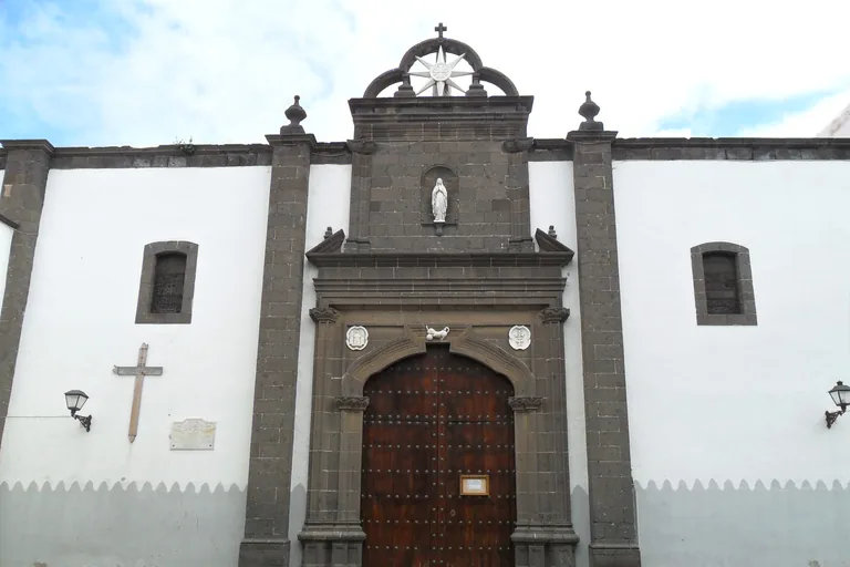 The white front of the church of Santo Domingo, a portal of dark grey stone round a tall arched wooden door, with a small white statue in a niche above and a star inside an openwork crown at the top.