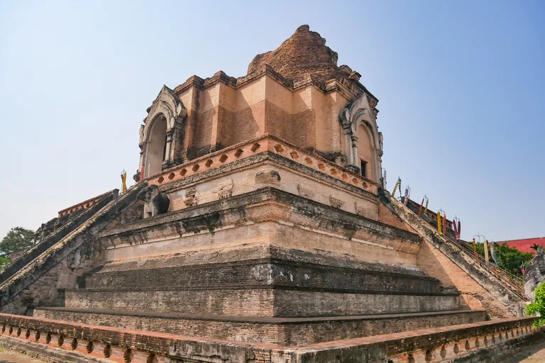 The great brick chedi of Wat Chedi Luang in Chiang Mai, its top broken away, with arched niches on its upper level, a stone elephant on a ledge and long stairs rising up its sides, under a clear blue sky.