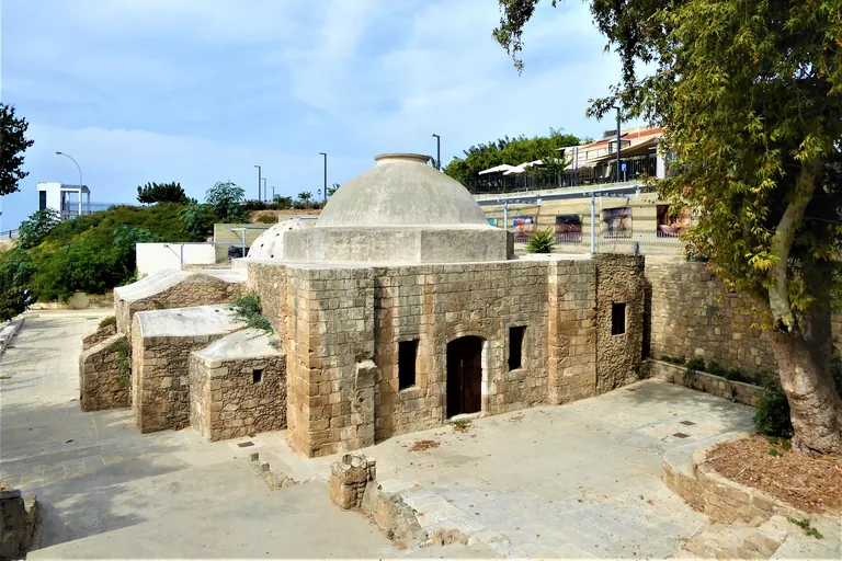 The Ottoman bath of Paphos seen from above, a square stone building with a plastered dome and a smaller dome pierced with small holes, low vaulted rooms beside it and a paved court around it, under a large tree.