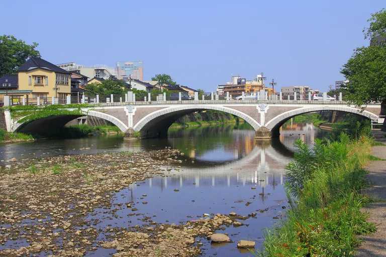 The Asanogawa Ohashi in Kanazawa, a pale concrete bridge of three arches with lamps along its railings, reflected in the shallow Asano river over a stony bed, with houses on the far bank and green weeds along a riverside path at the right.