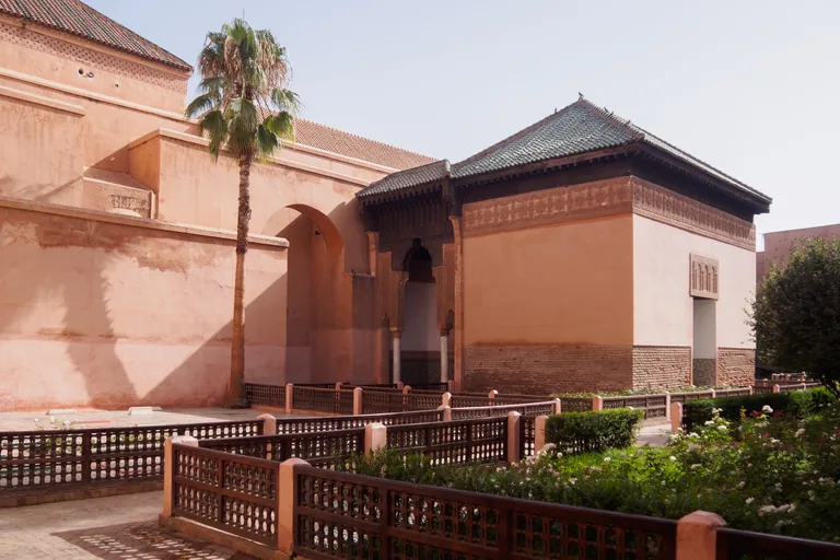 The pink walls and green tiled roofs of the Saadian Tombs above a garden of rose bushes enclosed by low wooden lattice fences, with a tall palm tree against the wall.