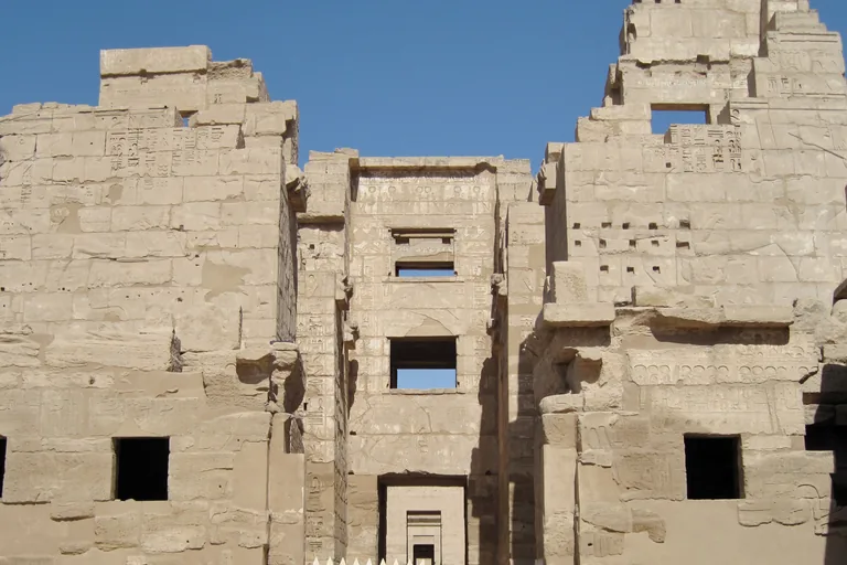 The two stone towers of the High Gate at Medinet Habu rising against a blue sky, with carved walls, the windows of the upper rooms between them and the passage leading through to further doorways.