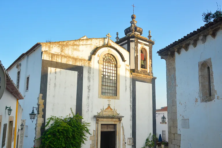 The whitewashed front of the Church of São Pedro, with dark trim, a tall barred window over the doorway and a bell tower with a rounded top.
