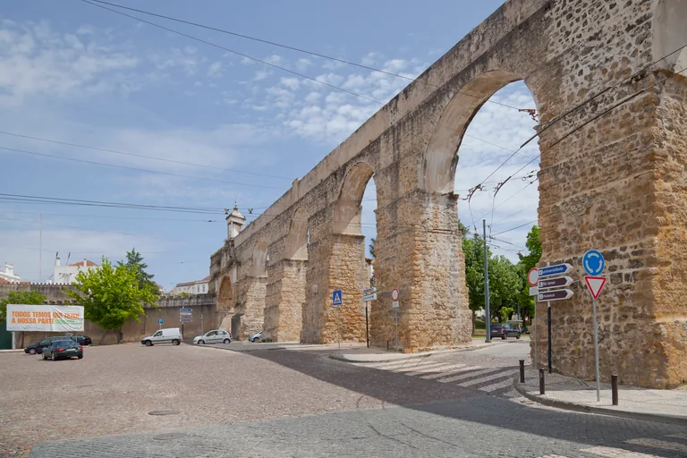 The tall stone arches of the São Sebastião Aqueduct crossing a cobbled road, with a small temple on the arch in the distance.