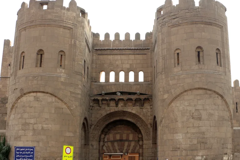 Bab al Futuh seen from the north, two round crenellated stone towers on either side of a deep arched gateway, with a row of small arched openings above the arch and the city wall continuing on both sides.