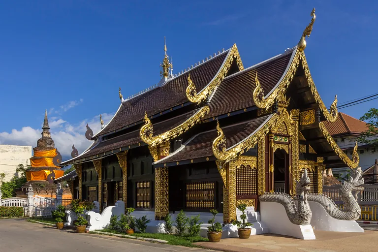 The dark wooden hall of Wat Inthakhin in Chiang Mai beside a quiet street, its tiered roofs edged in gold, a gilded porch with two white serpent figures on its steps, and an old brick chedi at the left under a deep blue sky.