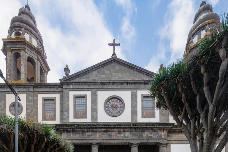 The upper front of La Laguna Cathedral, grey stone and white panels under a pediment and a cross, between two domed bell towers, with dragon trees at both sides and clouds in a blue sky.